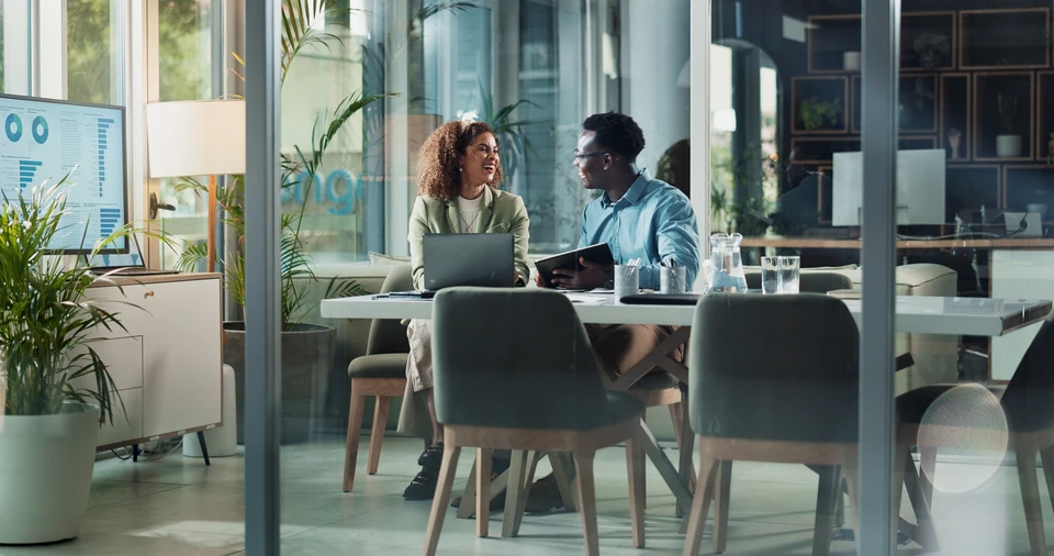 Deux professionnels collaborent dans un bureau moderne, une femme blonde en chemise bleue pointant vers un écran d'ordinateur tandis qu'un homme consulte des données